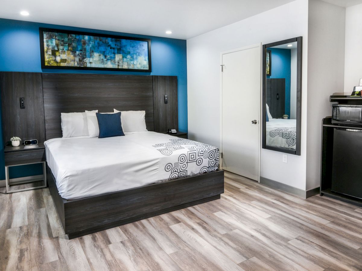 This image shows a modern hotel room with a large bed, artwork above the headboard, a mirror, and a mini-fridge and microwave corner set up.