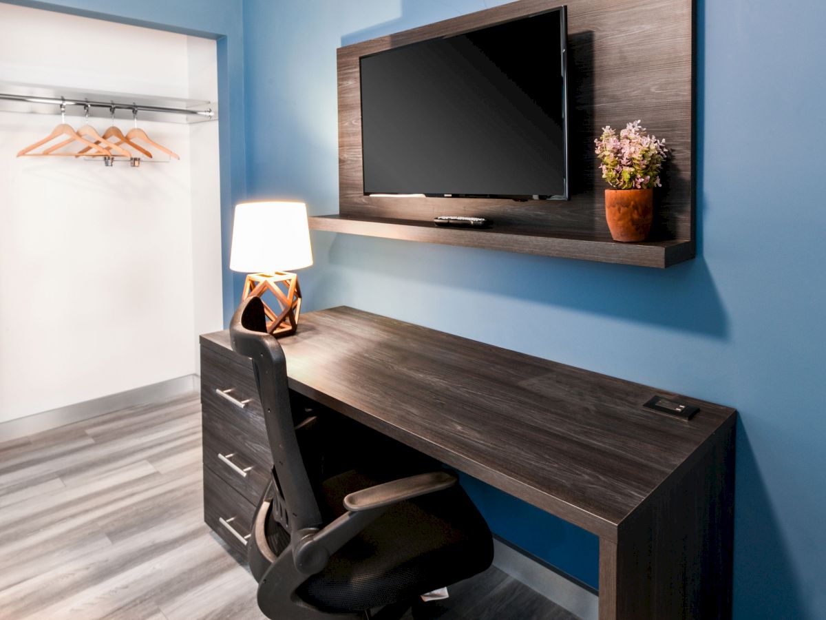 A modern office setup with a desk, chair, lamp, TV, and a plant on a shelf. There is an adjacent closet with hangers on the left side.