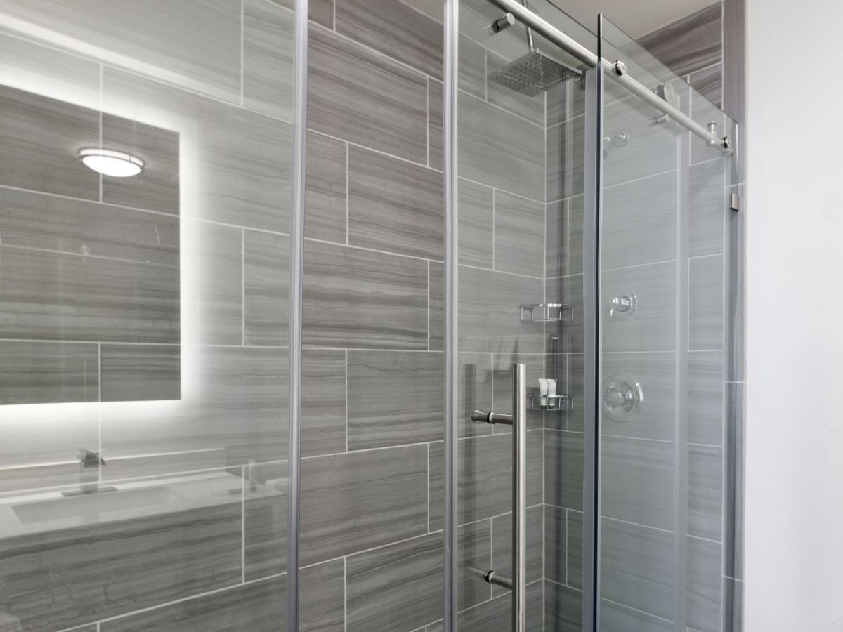 The image shows a modern bathroom with gray tile walls, a glass shower door with a metal handle, and a backlit mirror reflecting the design.