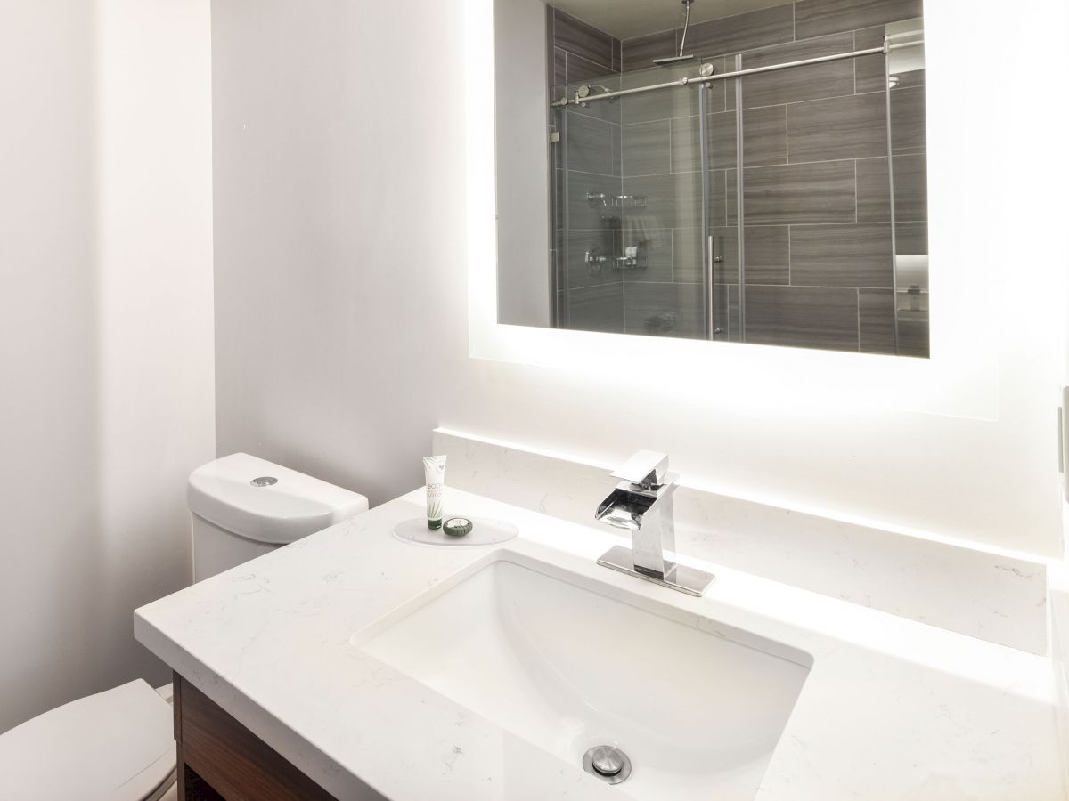 The image shows a modern bathroom with a sink, a toilet, and a large mirror with backlighting. The shower has glass sliding doors.