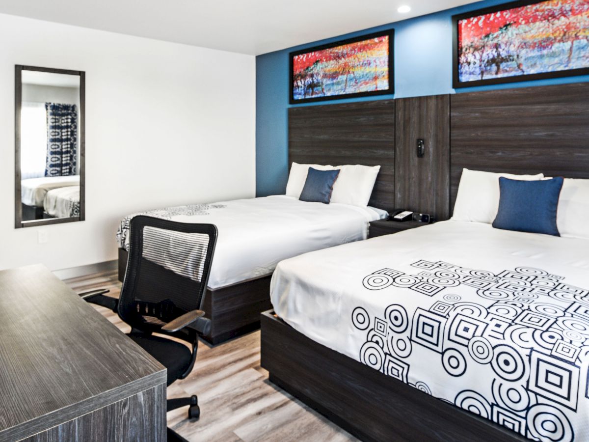 A modern hotel room with two double beds, wooden furnishings, abstract wall art, a desk, chair, and mirror.