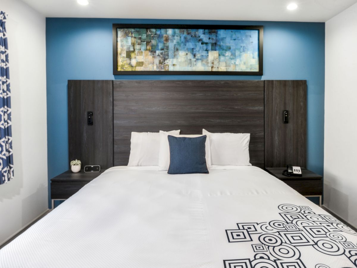 A modern bedroom with a large bed featuring white linens and geometric patterns, a blue accent wall, contemporary artwork, and bedside tables.