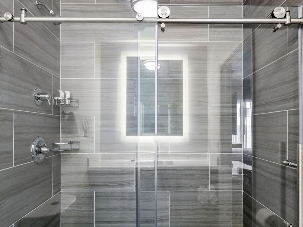 A modern shower with grey tiles, glass doors, and a backlit mirror on the wall. There is a showerhead, faucet, and shampoo holder on the left.