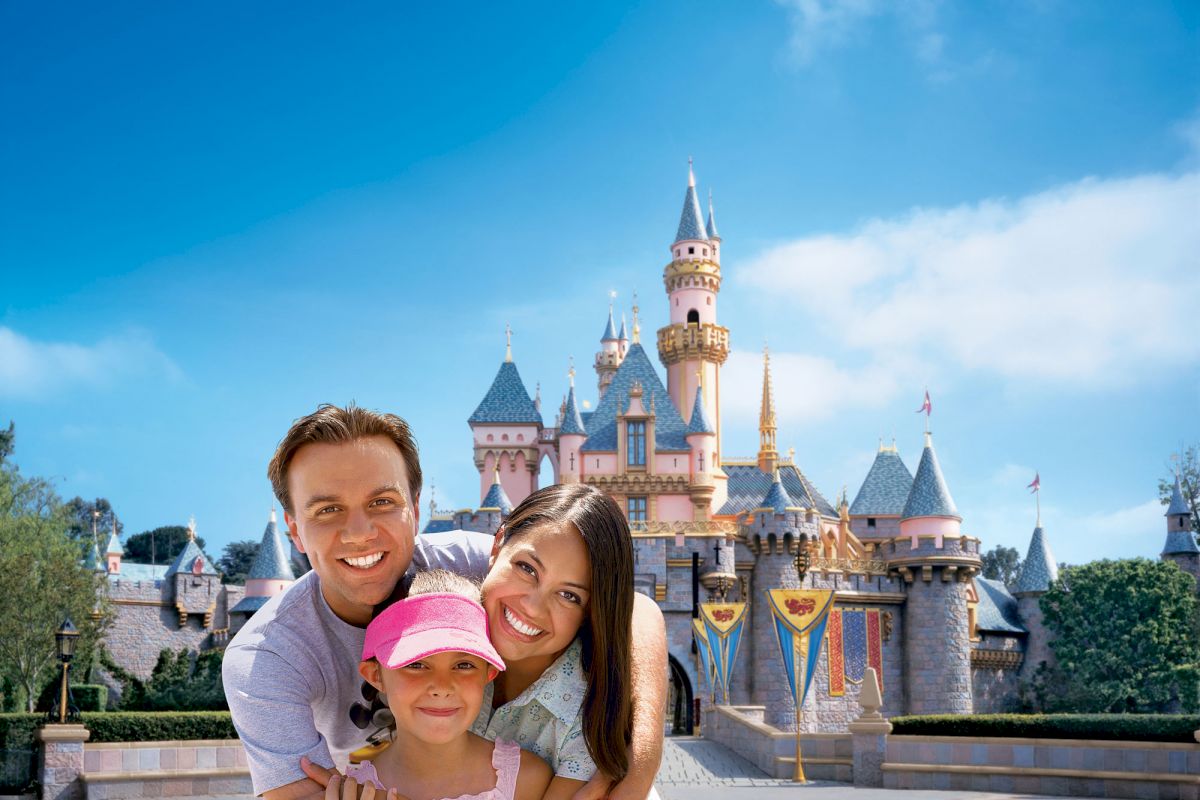 A happy family poses in front of a castle at an amusement park, with clear blue skies and cheerful expressions on their faces, creating a joyful scene.