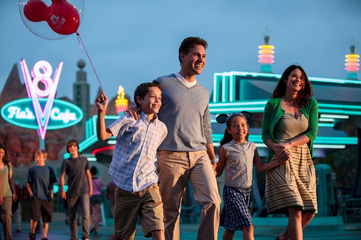 A family is walking together at an amusement or theme park, holding hands and smiling, with neon lights in the background and a child holding balloons.