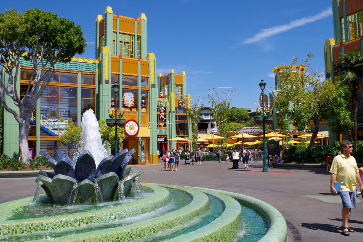 An outdoor scene in a park with a fountain, colorful buildings, trees, and people walking around on a sunny day.