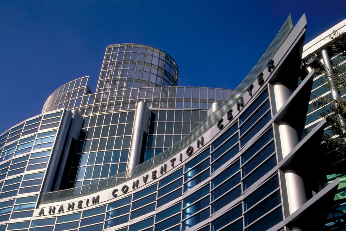 The image shows the exterior of the Anaheim Convention Center with its distinctive glass and metal architectural design, under a clear blue sky.