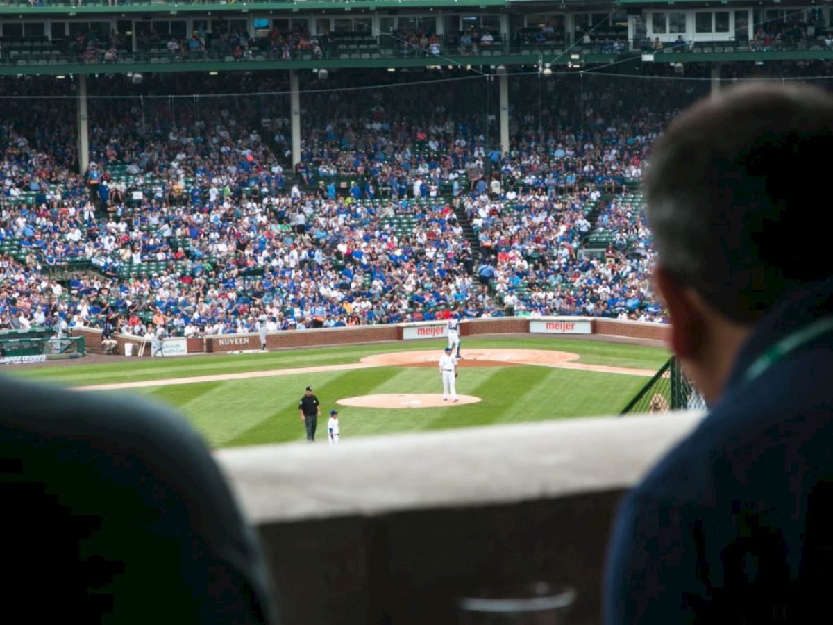 Two spectators view a baseball game from behind home plate, with the field and crowded stadium in the background.