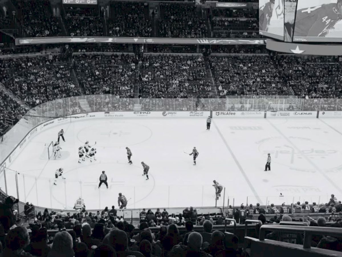 A large crowd watches an ice hockey game in an indoor arena, with players on the rink and a giant screen overhead.