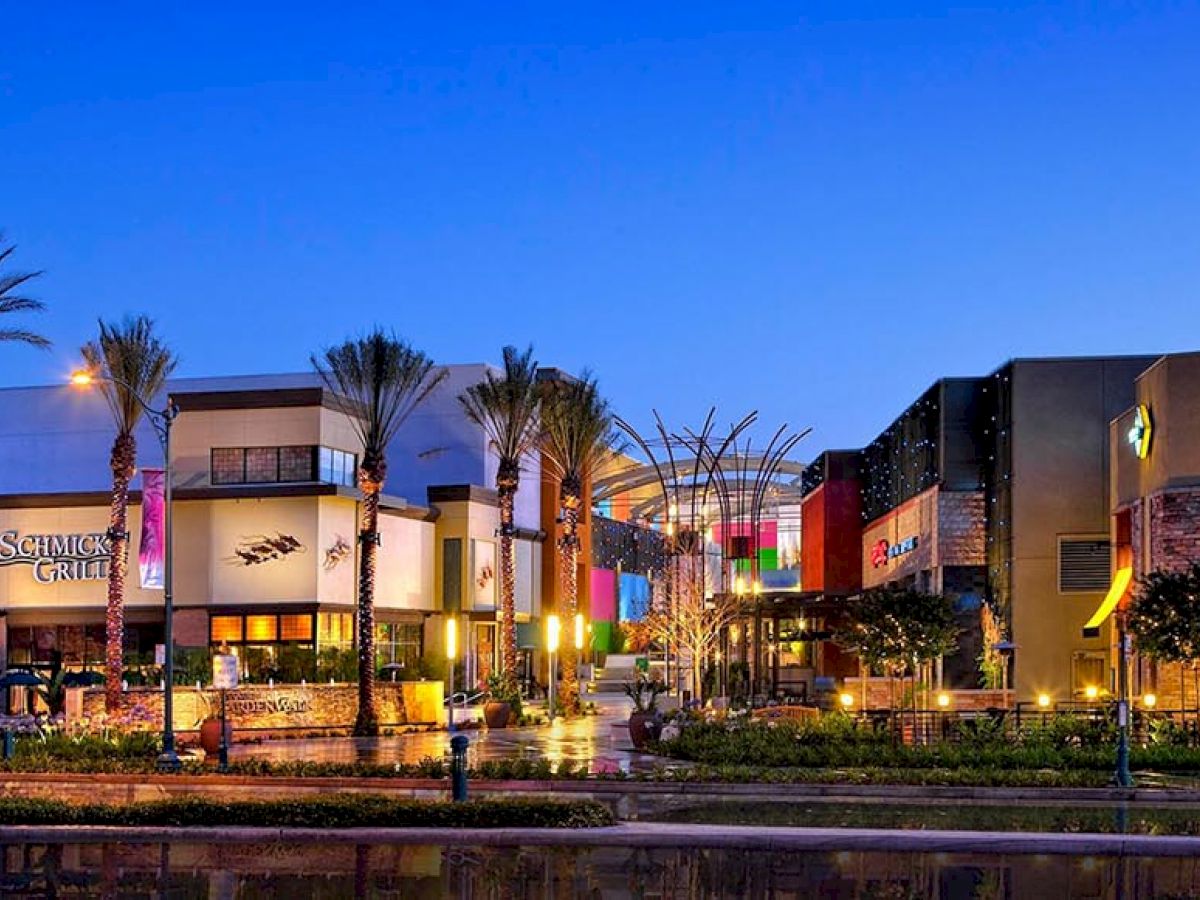 An outdoor shopping center with various stores, restaurants, palm trees, and colorful lights, captured in the evening under a clear sky.