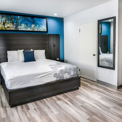 The image shows a modern hotel room with a bed, nightstands, wall art, a mirror, a mini fridge, microwave, coffee maker, and wooden flooring.