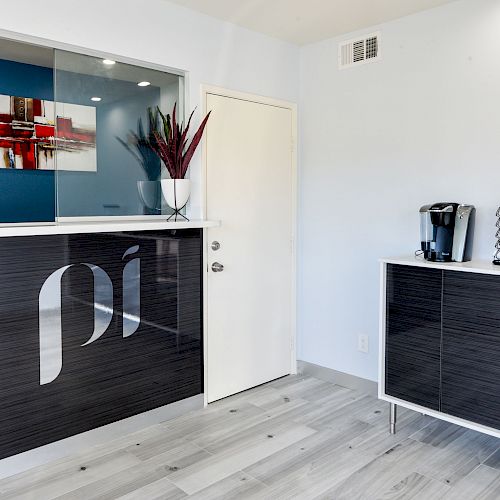 The image shows a modern office reception area with a front desk, wall art, a coffee station with cups, and a cabinet.