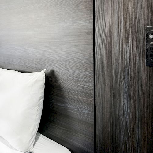 The image shows a modern, dark wood paneled wall with a digital lock or electronic control panel next to a bed with white pillows and bedding.