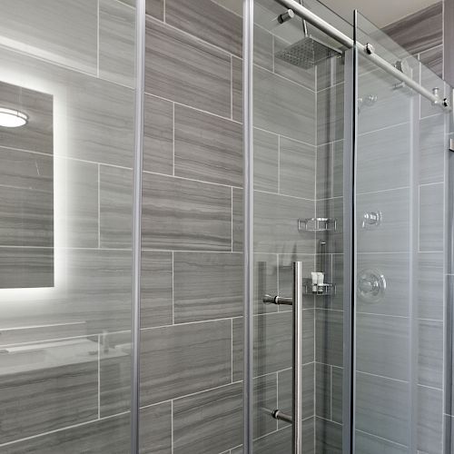 A modern bathroom features a glass-enclosed shower with gray tiles and a sleek sliding door. The mirror has a built-in light.