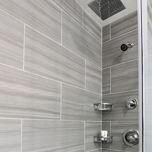 The image shows a modern shower with gray rectangular tiles, two showerheads, and a glass door. There are two metallic shelves holding toiletries.