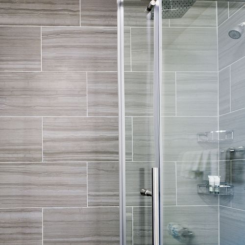 The image shows a modern glass-door shower with gray tiled walls, a wall-mounted showerhead, and built-in shelves holding toiletries and soap.