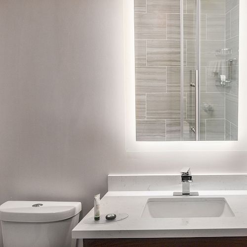 The image shows a modern bathroom with a toilet, a sink with a faucet, a large backlit mirror, and a shower area with glass doors.