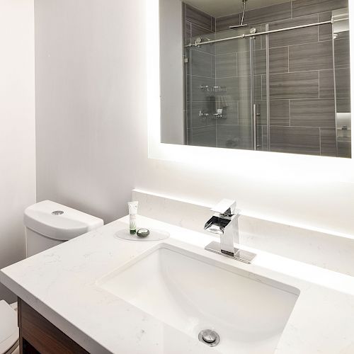 The image shows a modern bathroom with a white countertop, a sink, a faucet, a toilet, and a backlit mirror. The shower area has glass sliding doors.