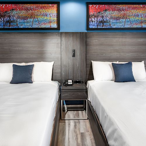 The image shows two neatly made beds with white linens and blue accent pillows, separated by a nightstand. There are colorful paintings above each bed.