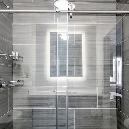 The image shows a modern, gray-tiled shower with a glass sliding door, built-in shelves, and a well-lit mirror inside the shower.