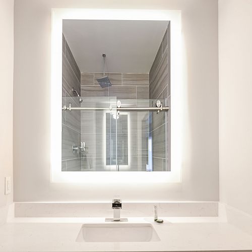 A modern bathroom with a backlit mirror, a sink with a faucet, and a glass-enclosed shower area in the reflection.