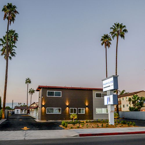 The image shows a modern two-story building surrounded by palm trees, with a lit-up sign near the entrance. The sky is clear during dusk.