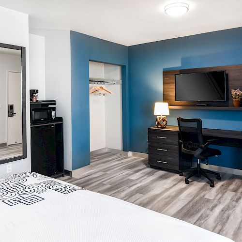 The image shows a modern hotel room with a bed, a desk with a chair and lamp, a wall-mounted TV, a mini-fridge, a closet, and a bathroom entrance.