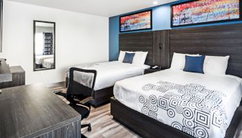 A modern hotel room with two beds, desk, chair, mirror, and colorful artwork above the beds. Simple, contemporary design and decor.