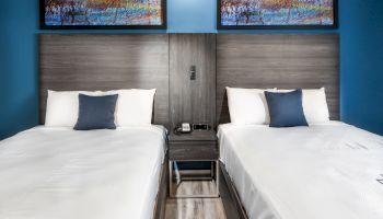 The image shows a hotel room with two neatly made beds, each with white linens and a dark blue pillow. Two colorful paintings are hung above the headboards.