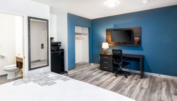 The image shows a modern hotel room with a bed, a TV on a wall, a desk and chair, a small kitchenette, a closet with hangers, and a bathroom.