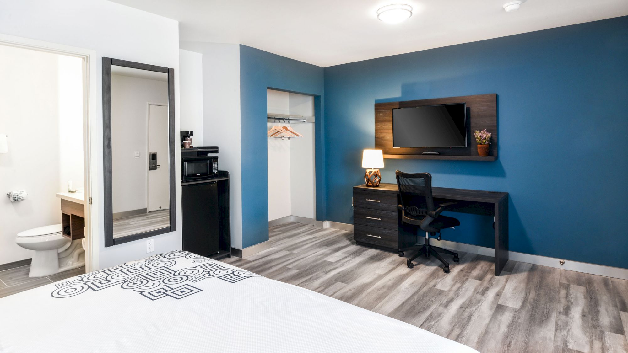 A modern hotel room with a bed, TV, desk, chair, lamp, closet, full-length mirror, and an adjacent bathroom featuring a toilet and sink is shown.