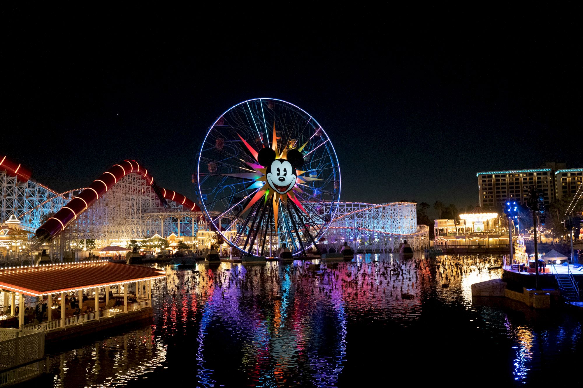 The image shows a night scene of a theme park with a large Ferris wheel featuring a cartoon face, lit up and reflecting in the water.