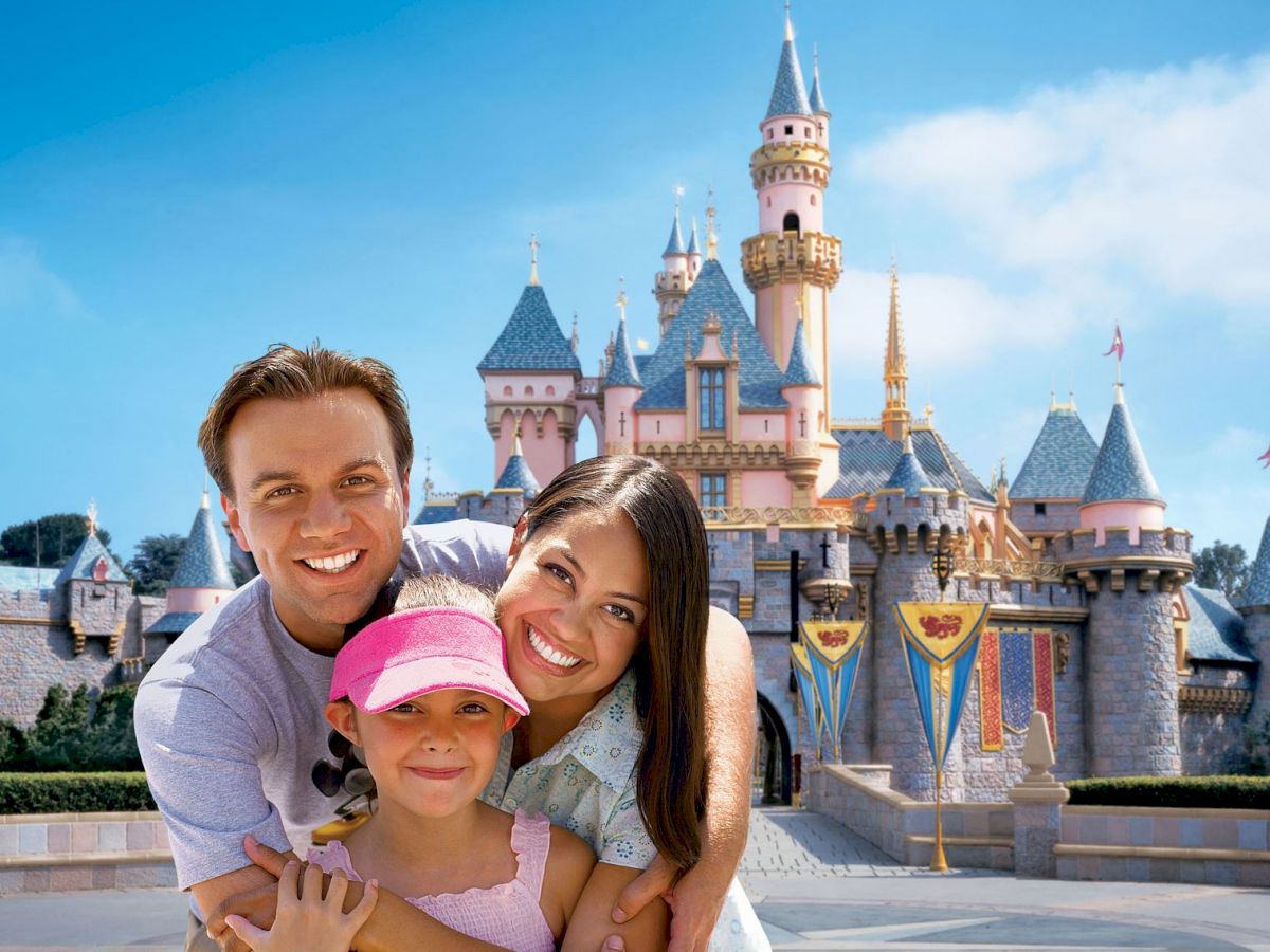 A family poses happily in front of a fairytale castle at an amusement park, with clear blue skies in the background.