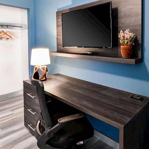 A modern workspace with a wooden desk, black chair, table lamp, potted plant, wall-mounted TV, and an open closet with wooden hangers.