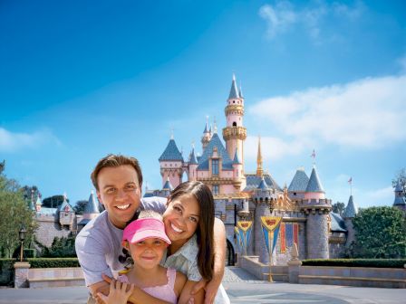 A smiling family posing in front of a castle at a theme park, with clear blue skies in the background.