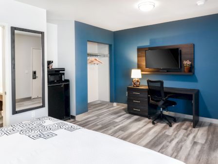 A modern hotel room features a bed, bathroom, mini-fridge, microwave, large mirror, desk with chair, and TV mounted on a blue accent wall.